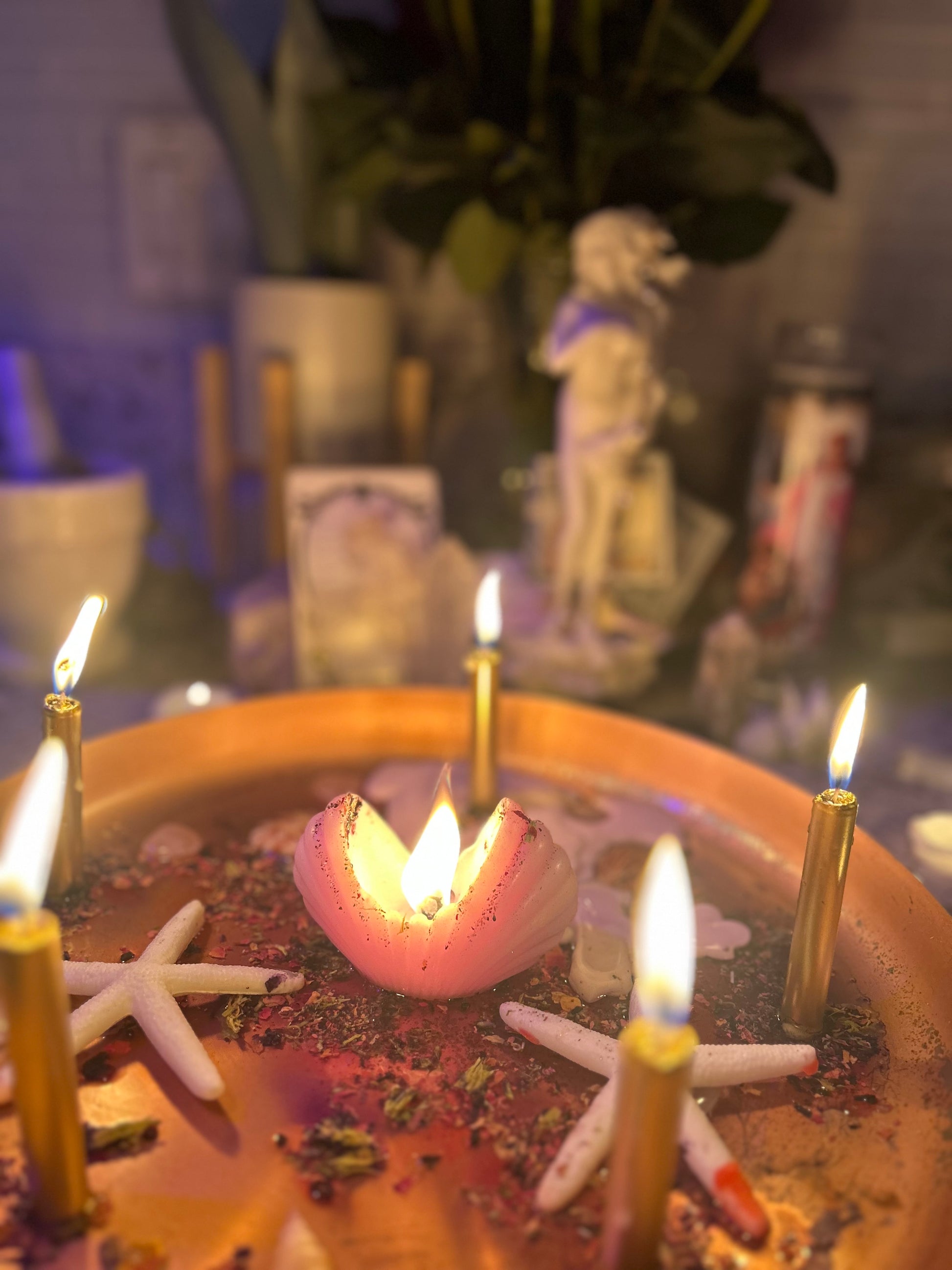 A mystical setup with lit candles, a central rose-colored candle, and various items that create an esoteric atmosphere, including a statue in the background.